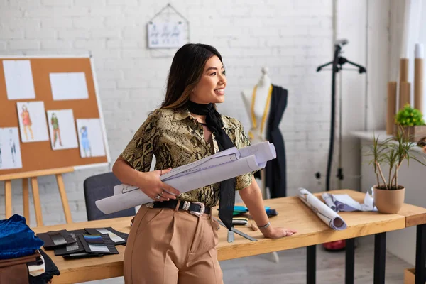 Радостный азиатский модельер с узорами шитья, смотрящий рядом с рабочим столом в личном ателье — стоковое фото