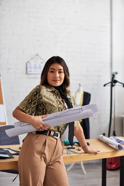 Sonriente asiático ropa diseñador con costura patrones mirando cámara cerca trabajo escritorio en propio atelier - foto de stock