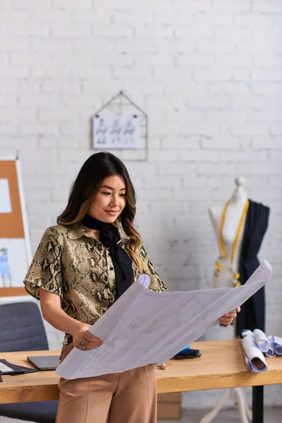 Diseñador asiático positivo mirando patrones de costura cerca de escritorio de trabajo en estudio de moda privada - foto de stock