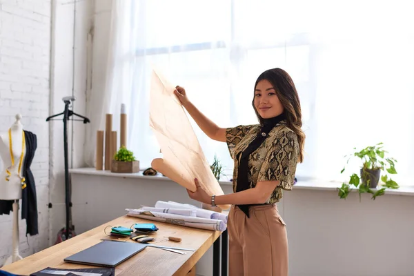 Young successful asian stylist with sewing patterns smiling at camera in modern fashion studio — Stock Photo