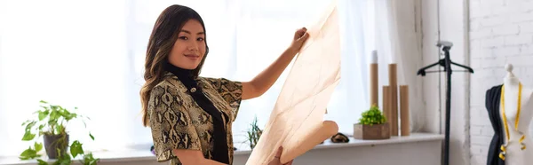 Joyful creative asian stylist with sewing patterns looking at camera in personal atelier, banner — Stock Photo