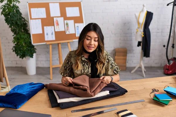Довольный азиатский персональный стилист, проверяющий качество образцов ткани на рабочем столе в собственном ателье — стоковое фото