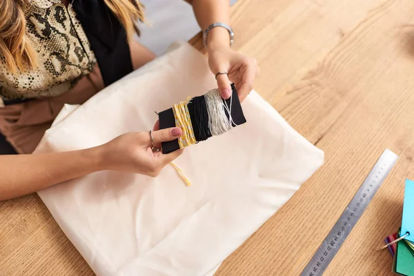 Vista recortada de modista sosteniendo hilos cerca de tela y suministros de costura en atelier personal - foto de stock