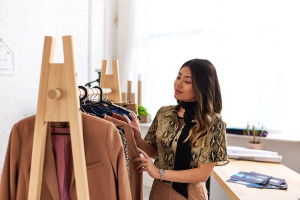 Young creative asian designer looking at fashionable bespoke garments on rack in personal atelier — Stock Photo
