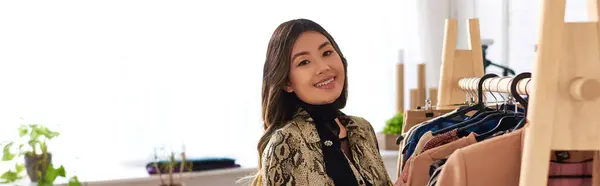 Счастливый азиатский персональный стилист смотрит в камеру возле стойки с модной одеждой в ателье, баннер — стоковое фото