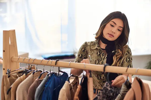 Attractive asian stylist looking at fashionable bespoke clothing on rack in personal studio — Stock Photo