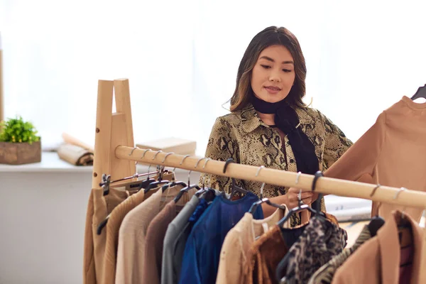 Ambitious asian stylist standing near trendy casual clothes on rack in modern fashion studio — Stock Photo