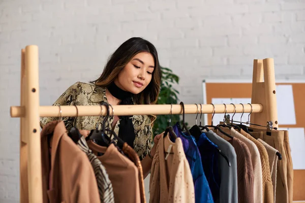 Молодой и талантливый азиатский стилист смотрит на модную одежду на заказ на стойке в частном ателье — стоковое фото