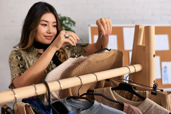 Zufriedene erfolgreiche asiatische Stylistin hält modische lässige Kleidungsstücke im persönlichen Modestudio — Stockfoto
