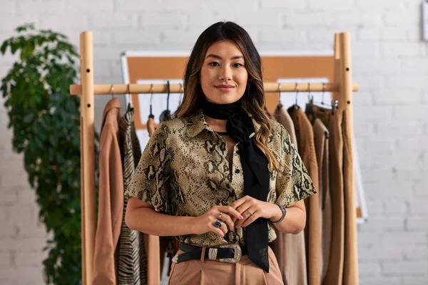 Young asian stylist in trendy casual attire smiling at camera near rack with clothes in atelier — Stock Photo