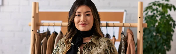 Fröhliche asiatische persönliche Stylistin blickt in die Kamera in der Nähe von Regal mit maßgeschneiderten Kleidungsstücken im Atelier, Banner — Stockfoto