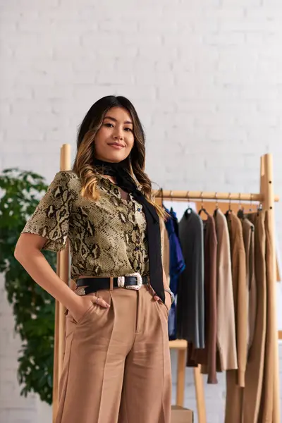 Joyful asian stylist in fashionable casual attire posing with hands in pockets in personal atelier — Stock Photo