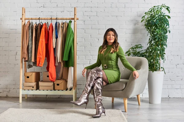Joven asiático mujer en moda casual atuendo posando en sillón cerca rack con ropa en propio atelier - foto de stock