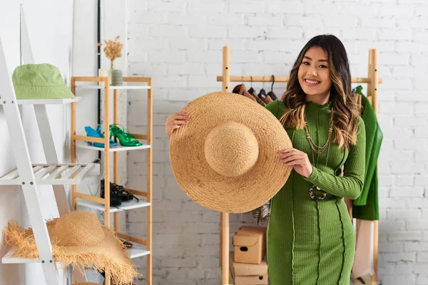 Joven y feliz asiático diseñador celebración de moda paja sombrero y sonriendo a la cámara en personal atelier - foto de stock