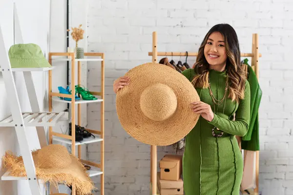 Young and glad asian designer holding trendy straw hat and smiling at camera in personal atelier — Stock Photo