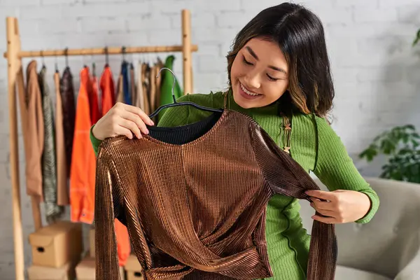 Complacido joven asiático diseñador de moda mirando elegante a medida blusa hecha en atelier personal - foto de stock