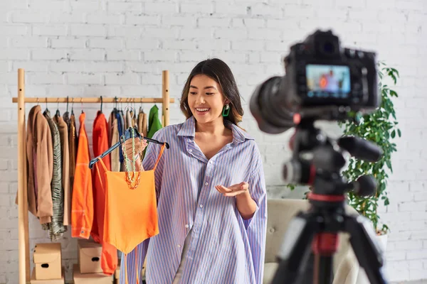 Улыбающийся азиатский дизайнер одежды, показывающий модный топ и аксессуары во время видео блога в ателье — стоковое фото