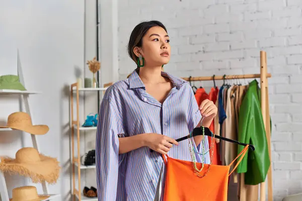 Pensativo asiático dressmaker con crop top y accesorios mirando lejos en atelier, negocio de la moda - foto de stock