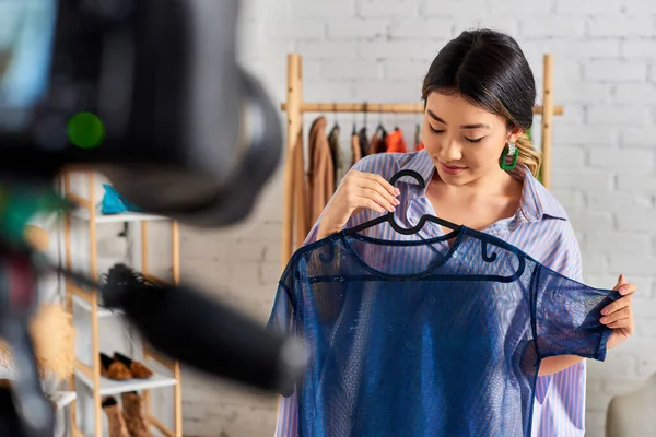 Молодая азиатская портниха с модной прозрачной блузкой возле размытой цифровой камеры в ателье — стоковое фото