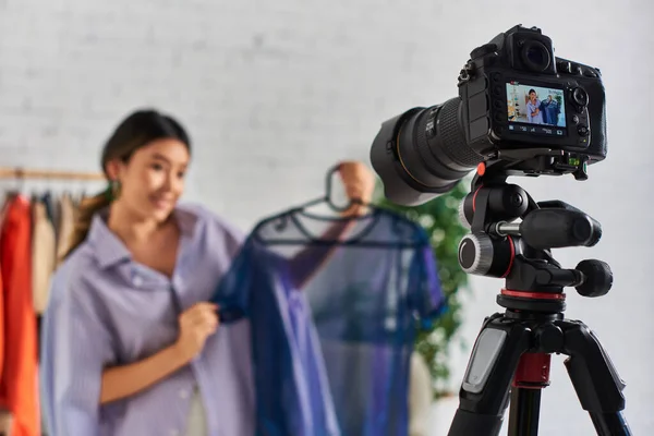Focus sur appareil photo numérique près de jeune styliste asiatique avec chemisier tendance en atelier, mode vlog — Photo de stock