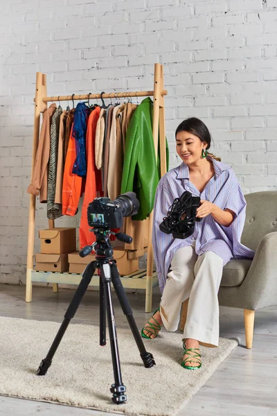 Joyful asian fashion designer holding trendy sandals during video blog in private clothes atelier — Stock Photo