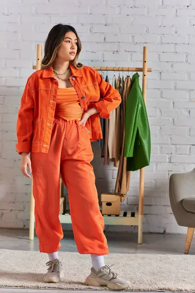 Trendy asian clothes designer in vibrant orange clothes posing with hands on hip in fashion studio — Stock Photo
