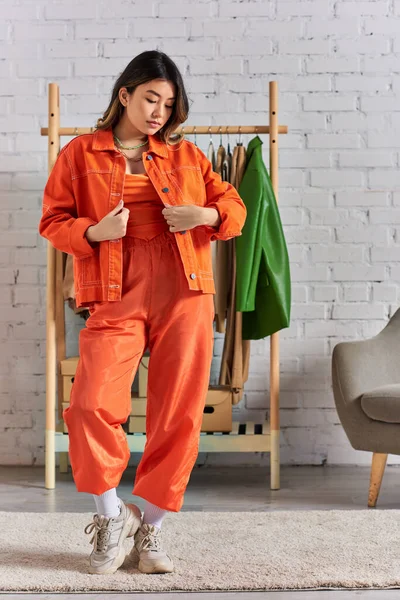 Экспрессивный азиатский стилист в модной оранжевой одежде позирует в личном ателье моды, малый бизнес — стоковое фото