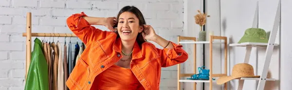 Encantado diseñador de moda asiático posando en ropa naranja brillante en el estudio, bandera horizontal - foto de stock