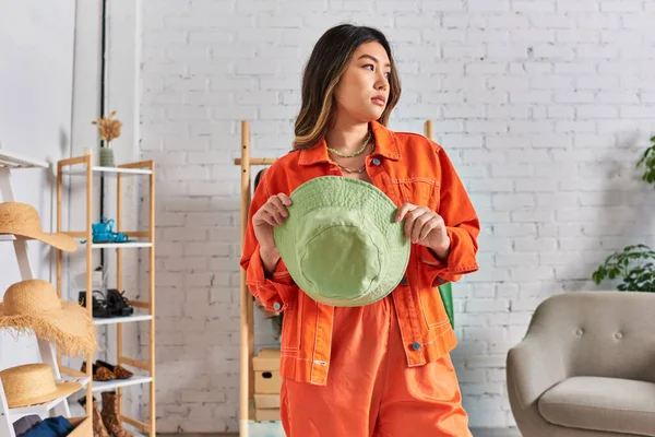 Thoughtful asian stylist in bright casual clothes holding panama hat and looking away in own atelier — Stock Photo