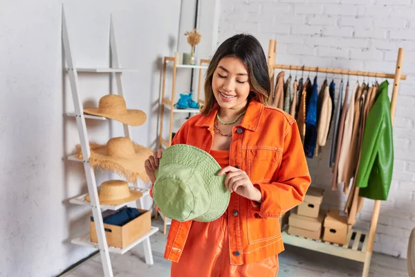 Pleased asian dressmaker in orange clothes holding panama hat in private atelier, small business — Stock Photo