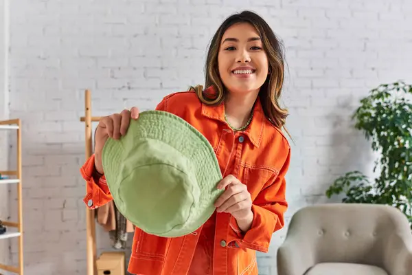 Excited asian stylist in orange clothes showing trendy panama hat at camera in fashion atelier — Stock Photo