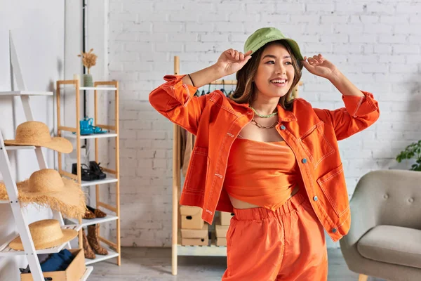 Восхитительный азиатский дизайнер в оранжевой одежде в панамской шляпе и глядя в собственный ателье — стоковое фото