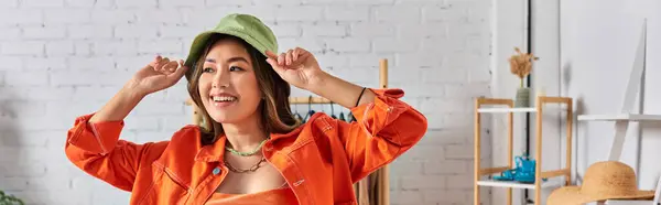 Happy asian stylist in orange attire wearing panama hat in clothes atelier, fashion business, banner — Stock Photo