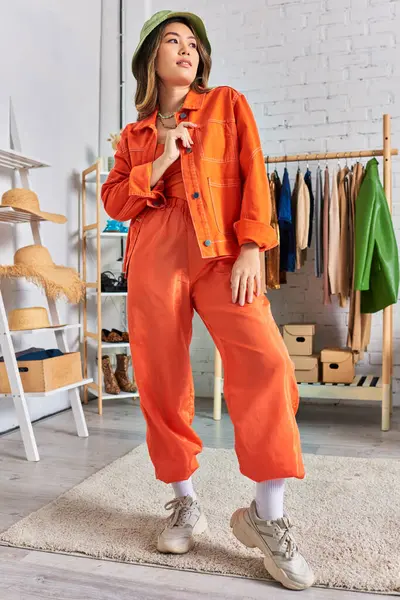 Confiante asiático designer em roupas laranja e chapéu panama posando no atelier pessoal moderno — Fotografia de Stock