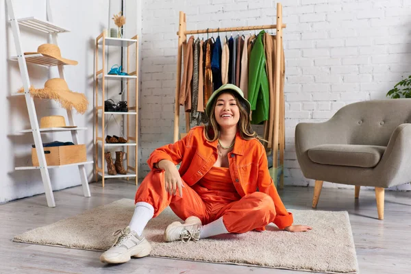Sonriente asiático estilista en brillante ropa y panama sombrero sentado en piso en personal moda atelier - foto de stock