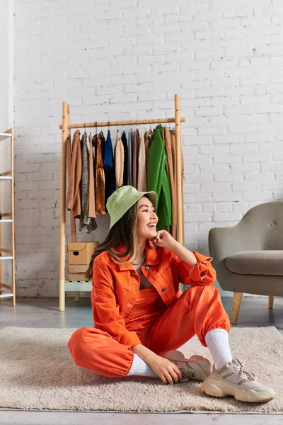 Alegre asiático diseñador en naranja ropa y panama sombrero sentado en piso en privado moda atelier - foto de stock