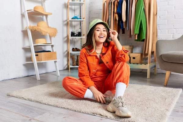 Carefree asian stylist in bright clothes and panama hat sitting on floor in personal fashion atelier — Stock Photo