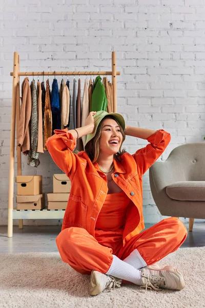 Overjoyed asiático costureira no panama chapéu e roupas brilhantes sentado no chão no atelier pessoal — Fotografia de Stock