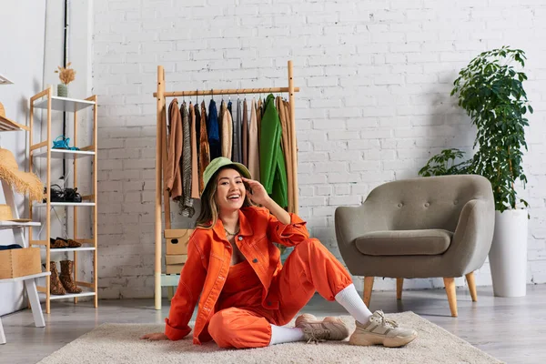 Happy asian stylist in orange clothes and panama hat sitting on floor in atelier, small business — Stock Photo