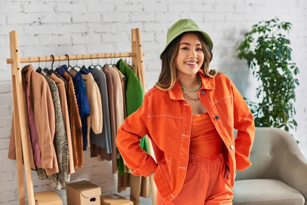 Successful asian stylist in orange attire and panama hat looking at camera in own fashion studio — Stock Photo