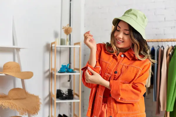 Joyful asian dressmaker trendy orange clothes and panama hat in personal atelier, small business — Stock Photo