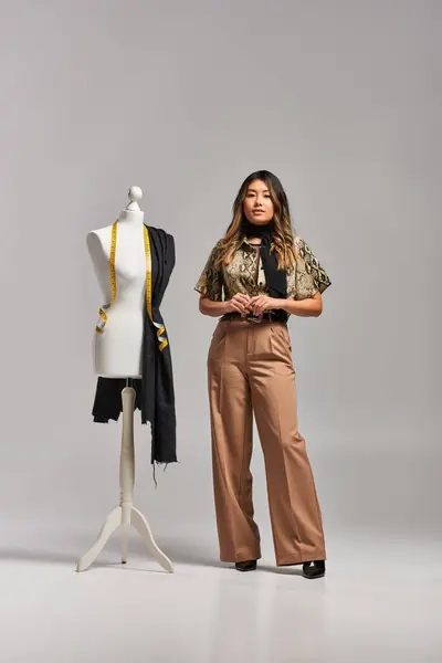 Trendy asian fashion designer posing near mannequin with measuring tape and fabric on grey backdrop — Stock Photo
