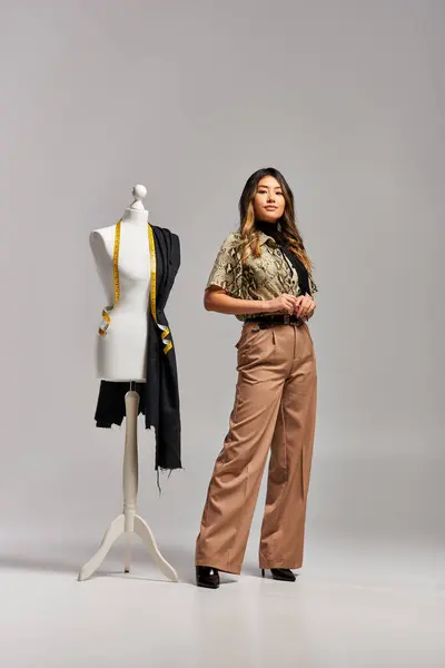 Confident asian stylist looking at camera near mannequin with measuring tape and fabric on grey — Stock Photo