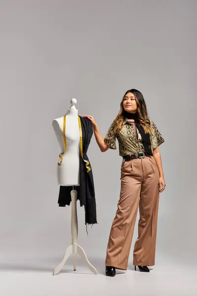 Exitoso diseñador asiático de moda mirando cerca maniquí con tela y cinta métrica en gris - foto de stock