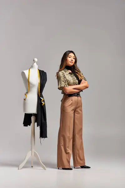 Designer asiático criativo com braços dobrados perto de manequim com tecido e fita métrica em cinza — Fotografia de Stock