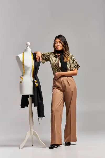 Happy trendy asian stylist posing near mannequin with fabric and measuring tape on grey backdrop — Stock Photo