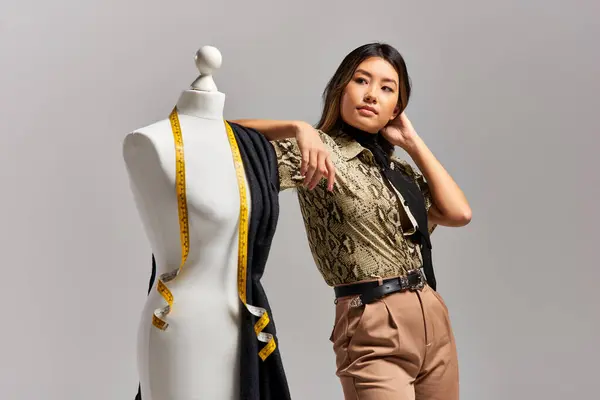 Творческий азиатский дизайнер смотрит в сторону рядом с манекеном с тканью и измерительной лентой на сером — стоковое фото