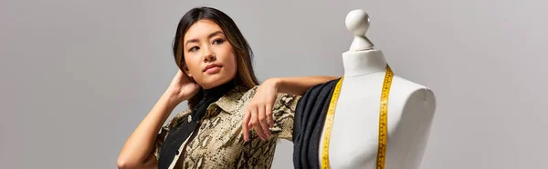 Modern asian stylist looking away near mannequin with fabric and measuring tape on grey, banner — Stock Photo