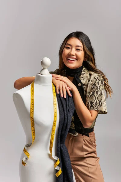 Веселый азиатский стилист смотрит на камеру рядом с манекеном с тканью и измерительной лентой на сером — стоковое фото