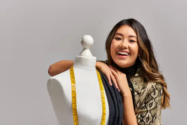 Excited asian stylist smiling at camera near mannequin with fabric and measuring tape on grey — Stock Photo